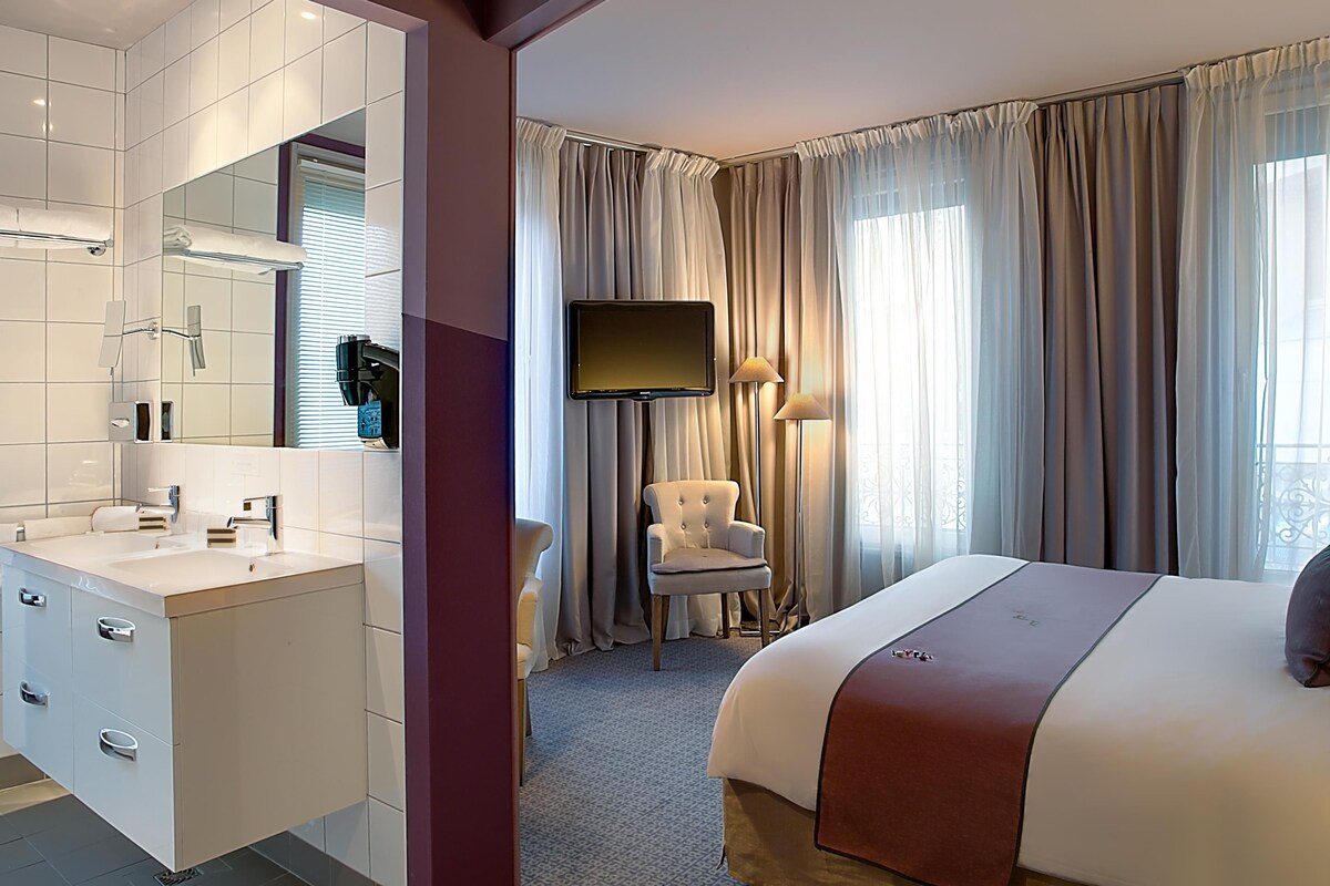 A hotel room with a large bed, a vanity with a mirror, a flat-screen TV, a chair, and a window with curtains.