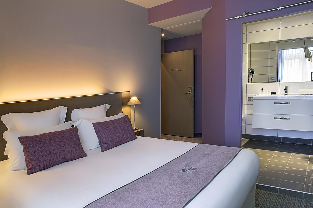 A modern hotel room with a large bed, a bedside table with a lamp, a bathroom with a mirror, and a door labeled "Cont. Lounge".