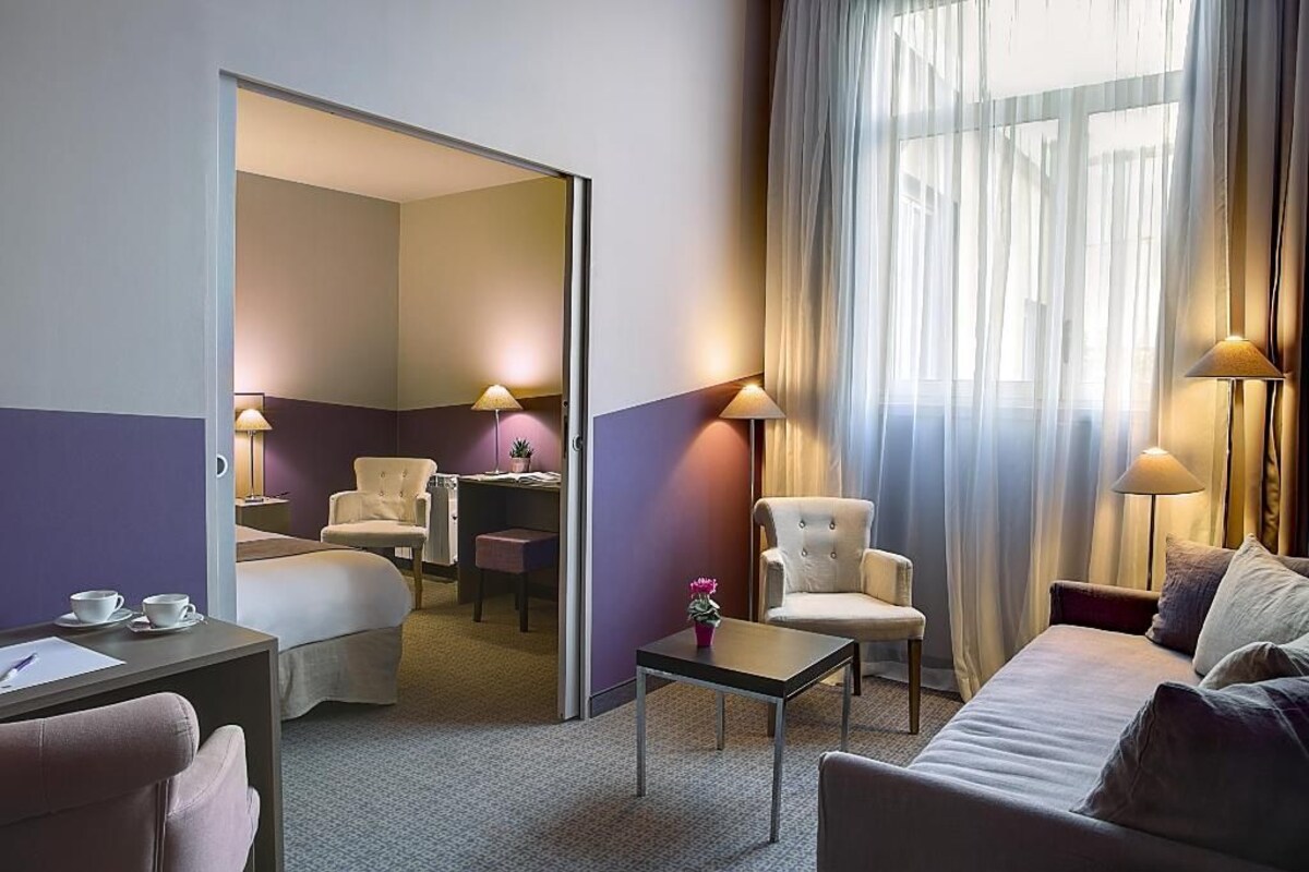 Une chambre d’hôtel avec un lit, un bureau, une chaise, un canapé et une grande fenêtre avec des rideaux.