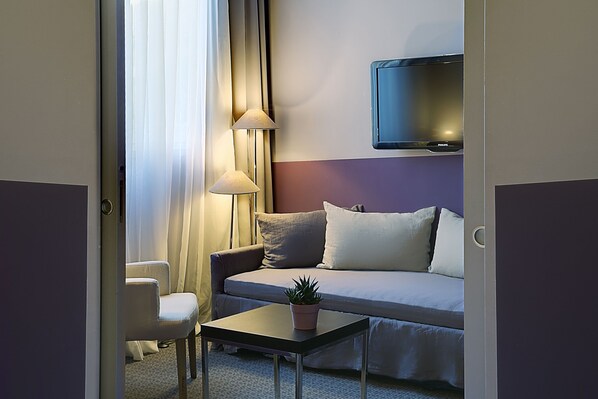 Une chambre d’hôtel avec un canapé, une petite table sur laquelle est posée une plante, une chaise, un téléviseur fixé au mur et une fenêtre avec des rideaux.