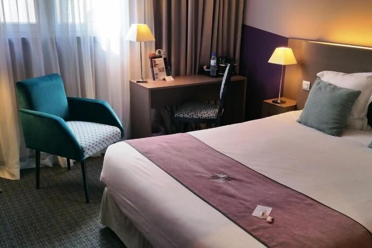 A hotel room with a bed, two pillows, a desk with a chair, a lamp, and a window with curtains.