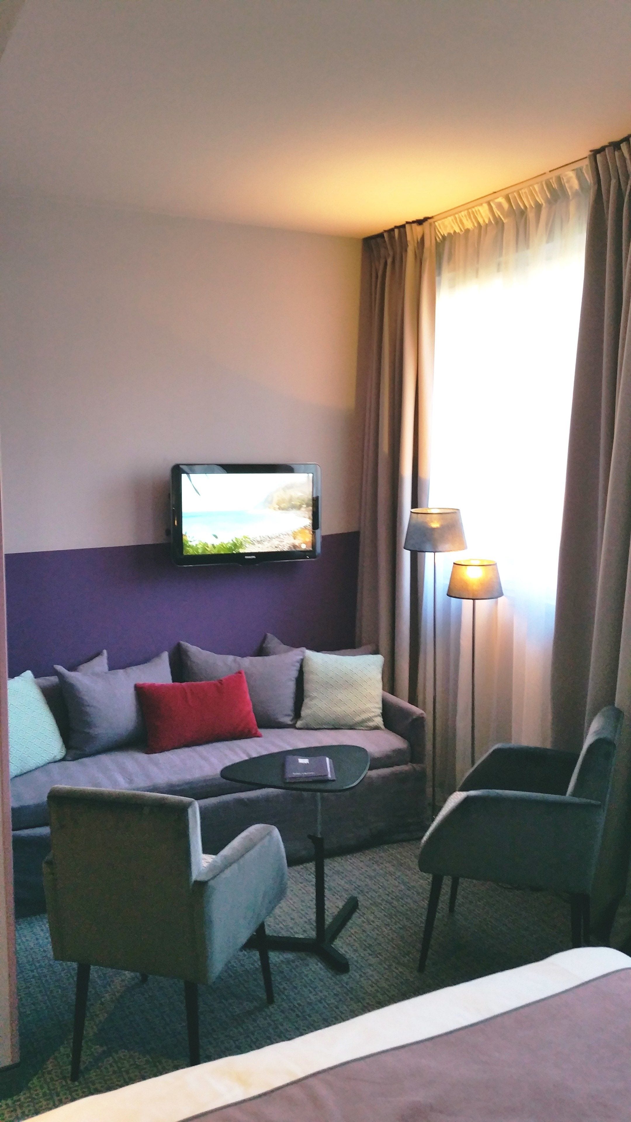 A hotel room with a sofa, two armchairs, a small coffee table, a television mounted on the wall, and a lamp.