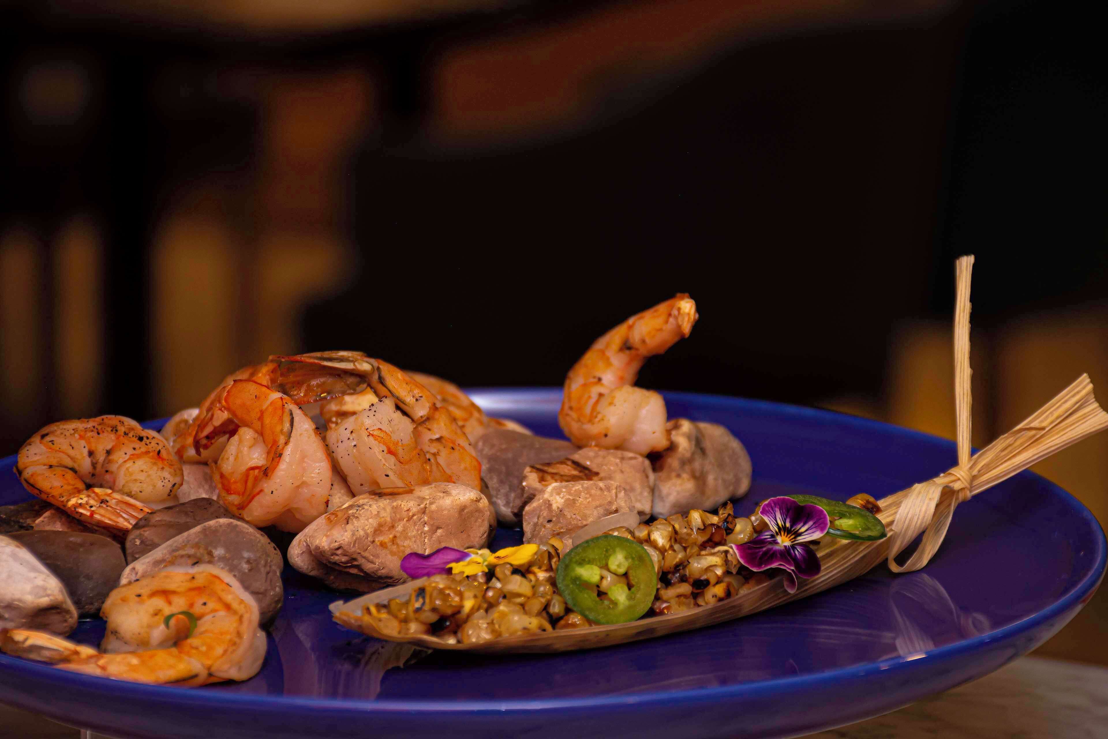 Un plato con diversos mariscos, como camarones, vieiras y un cangrejo, acompañado de una cucharada de maíz y una rodaja de jalapeño.