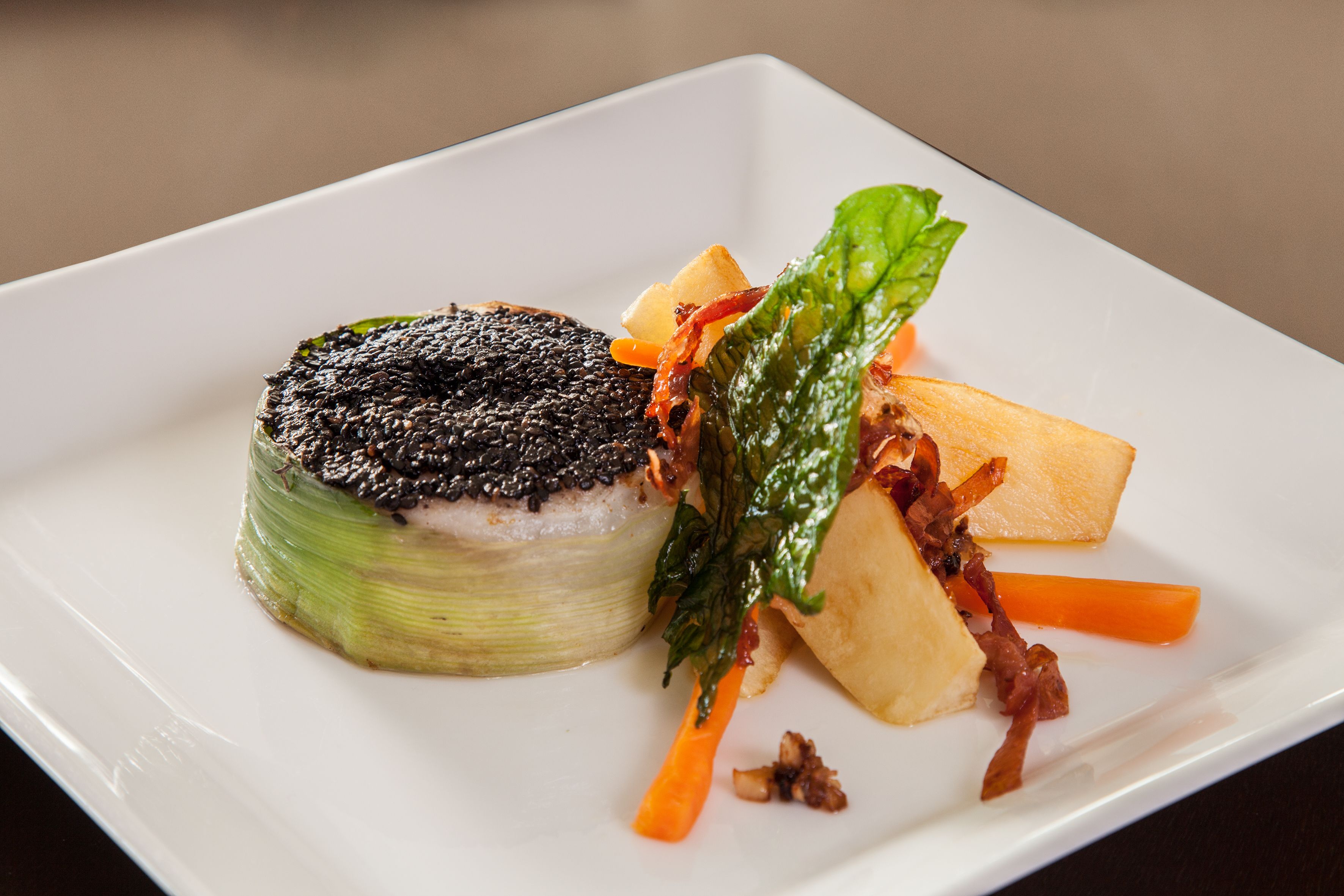 A square white plate with a round, layered food item topped with black sesame seeds, accompanied by a piece of tofu and a few carrot sticks.