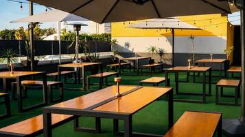 An outdoor seating area with multiple tables and umbrellas, surrounded by greenery and a yellow wall.
