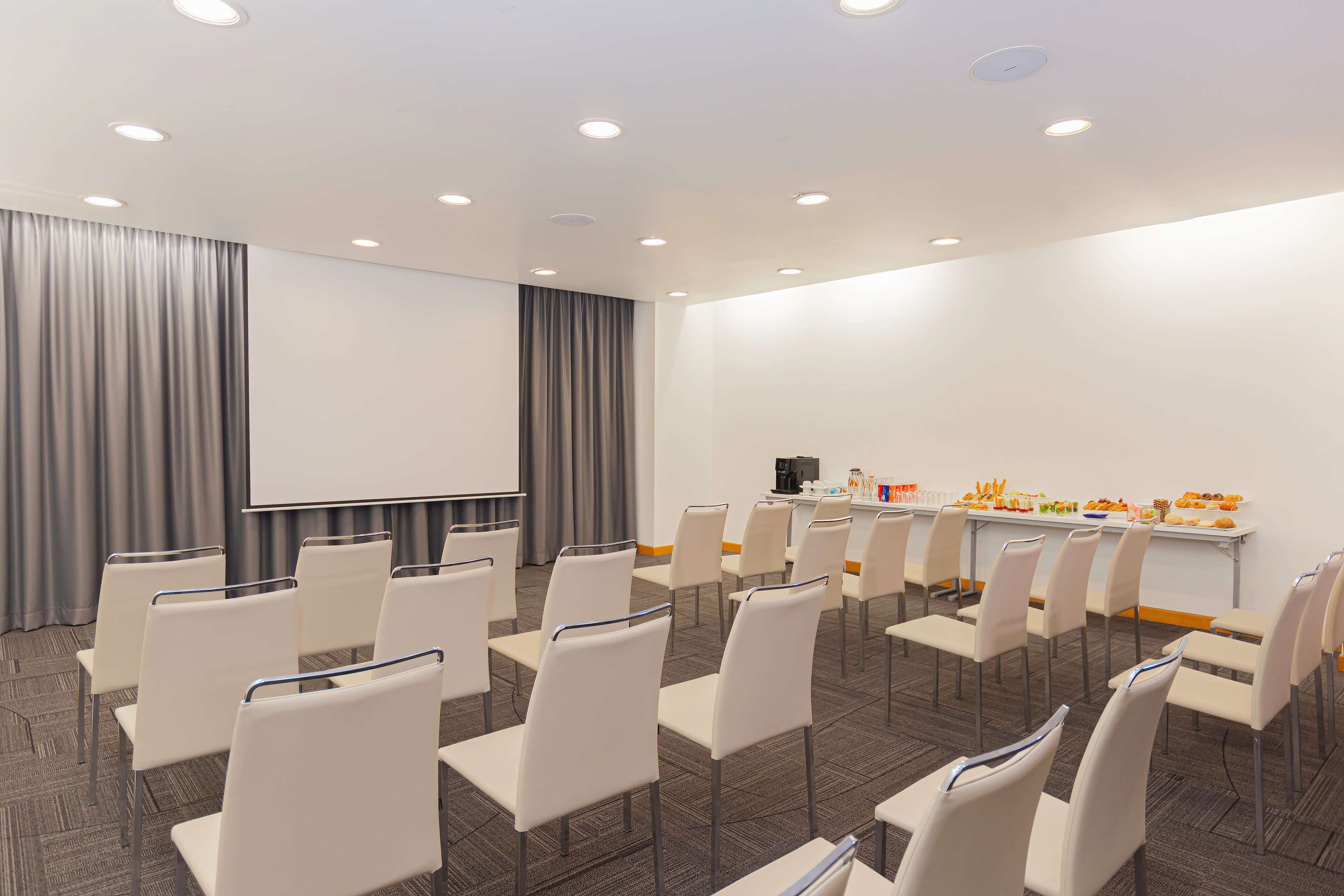 Ein Konferenzraum mit Reihen weißer Stühle, einer Projektionsleinwand und einem Buffet mit Speisen.