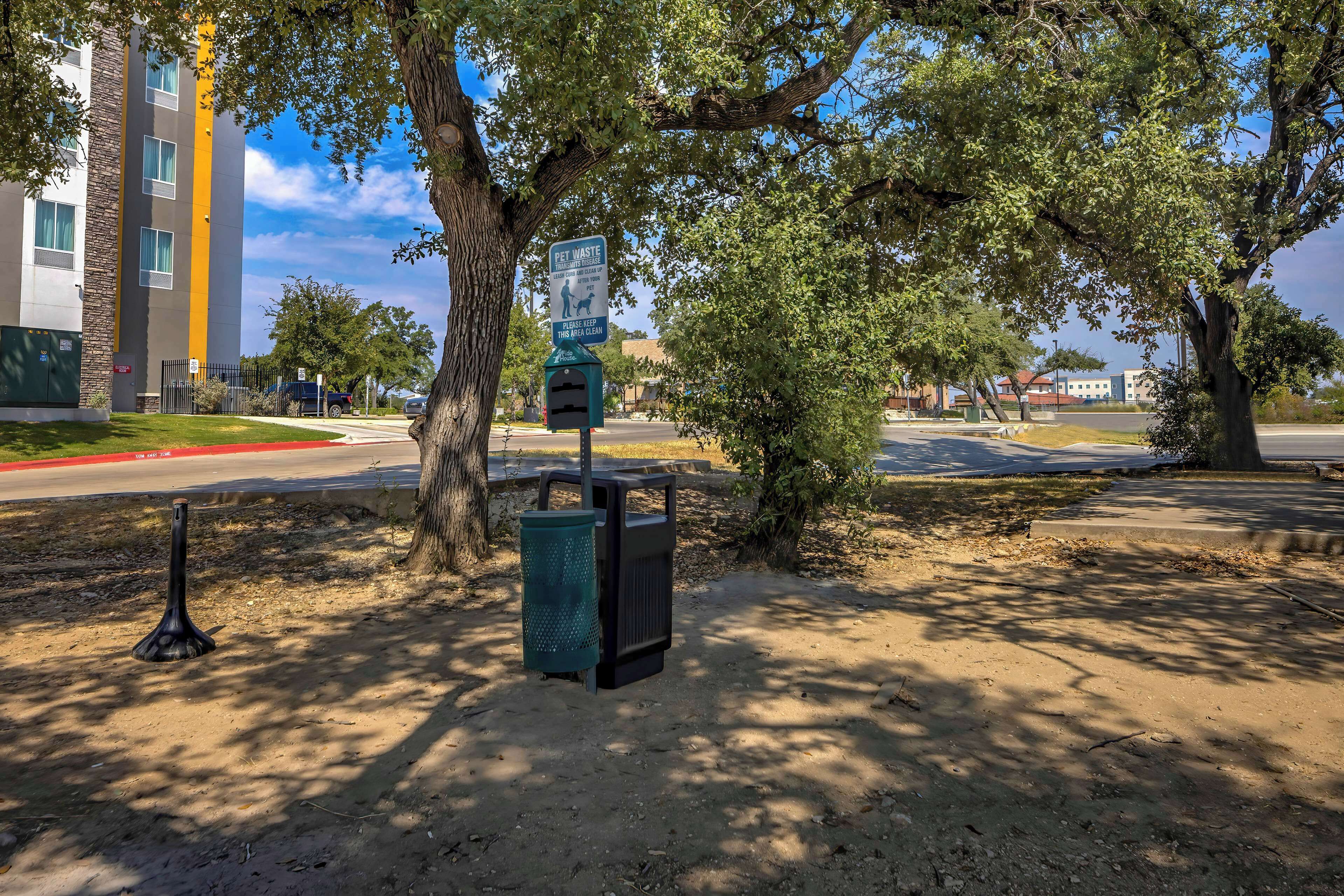 A pet waste station with a trash bin and a recycling bin, located in a shaded area with trees.