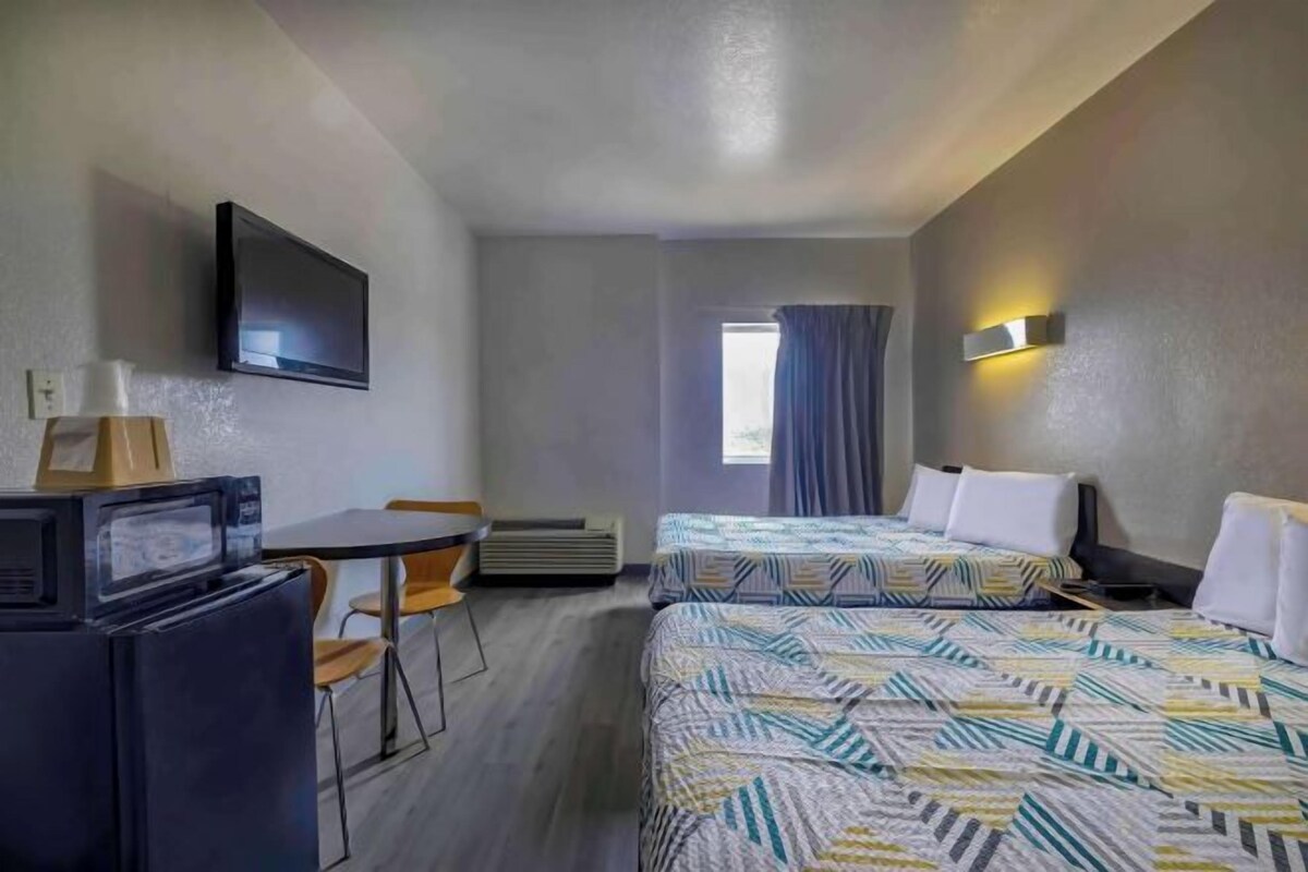 A hotel room with two beds, a desk with a computer, a flat-screen TV, and a window with curtains.