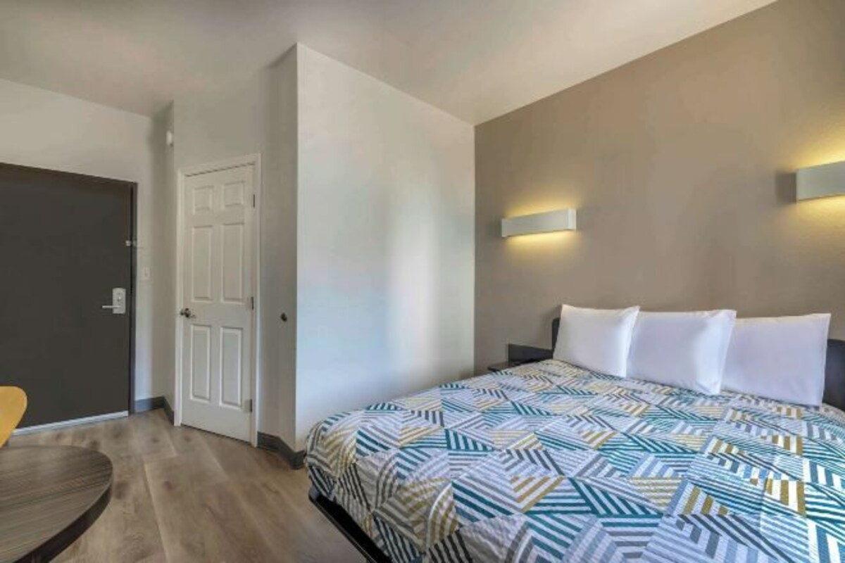 Quarto de hotel com cama, mesa de cabeceira, luminária na parede e porta.