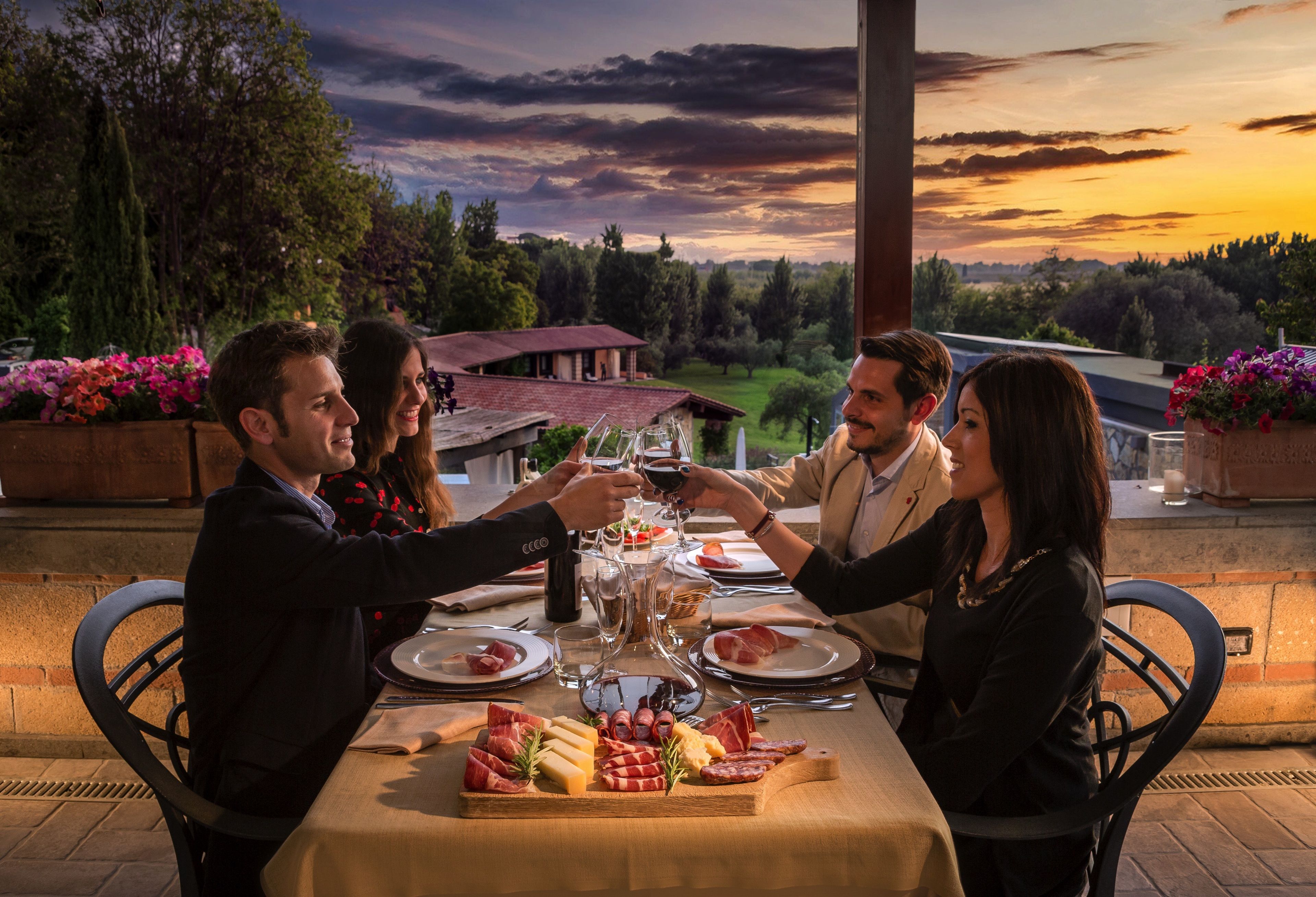 Un grupo de personas brindando en una mesa con una variedad de quesos y embutidos, con una vista panorámica al atardecer.