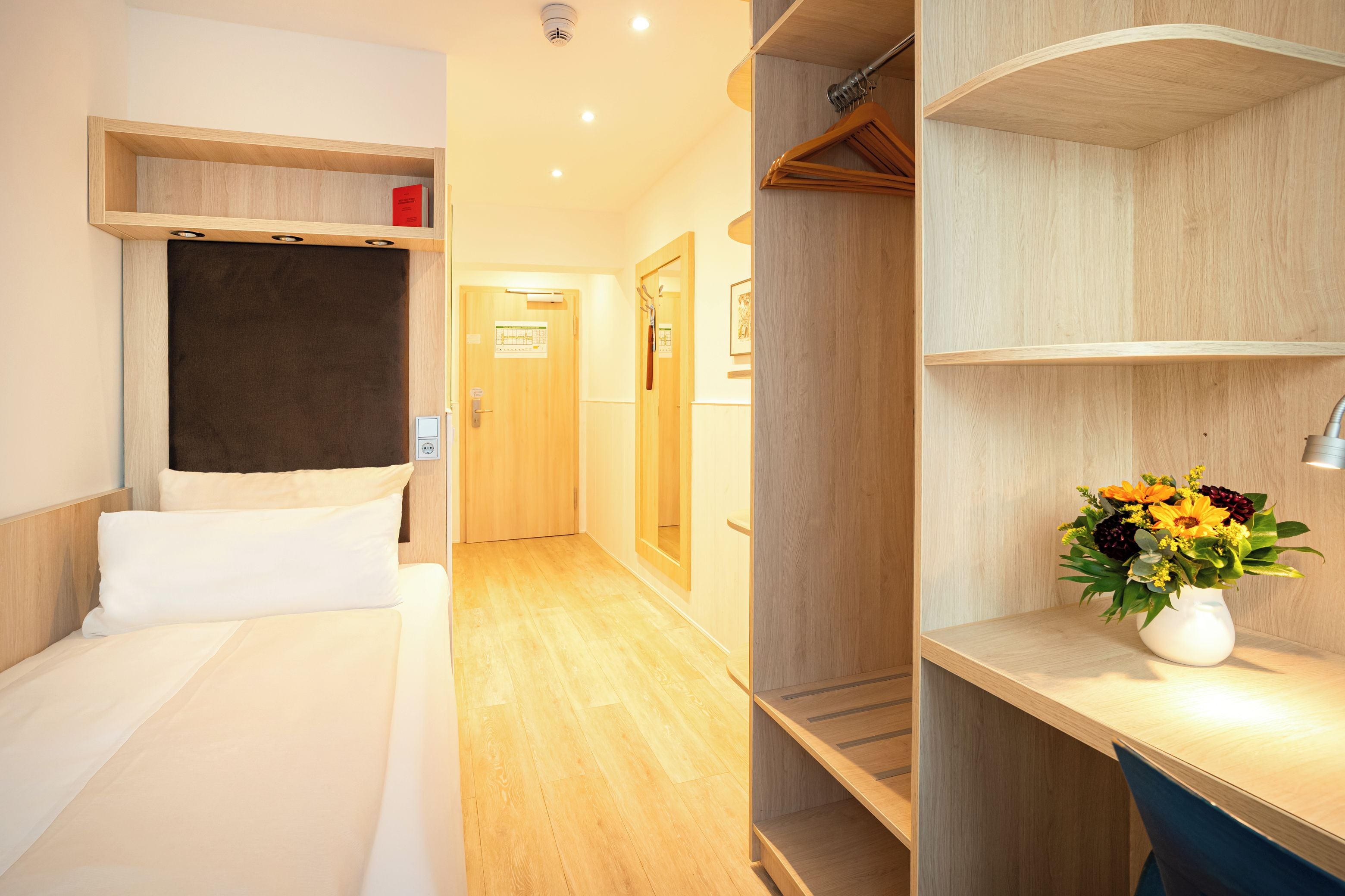 Una habitación de hotel con una cama, un armario de madera, un escritorio con lámpara y un jarrón de flores.