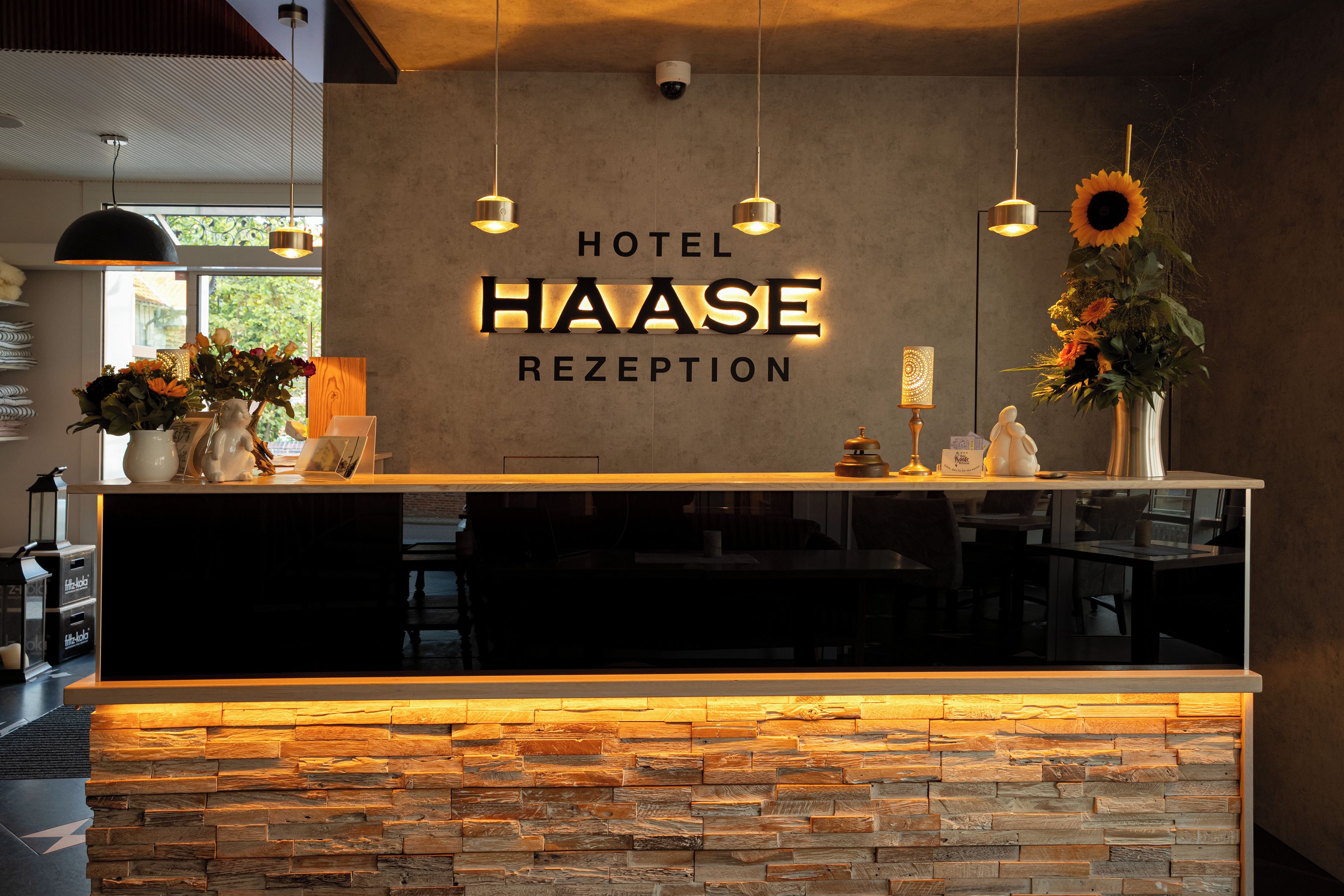 A hotel reception area with a stone wall, a sign reading 'HOTEL HAASE RECEPTION', and a vase with flowers.