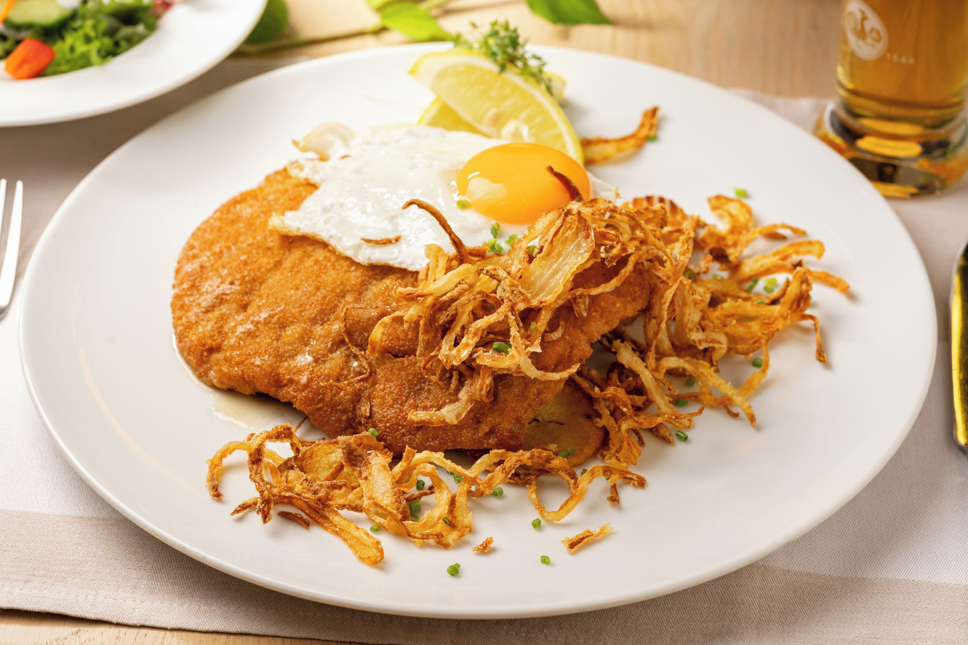 Un plato con una milanesa, cebollas fritas, un huevo frito y una rodaja de limón sobre un plato blanco.