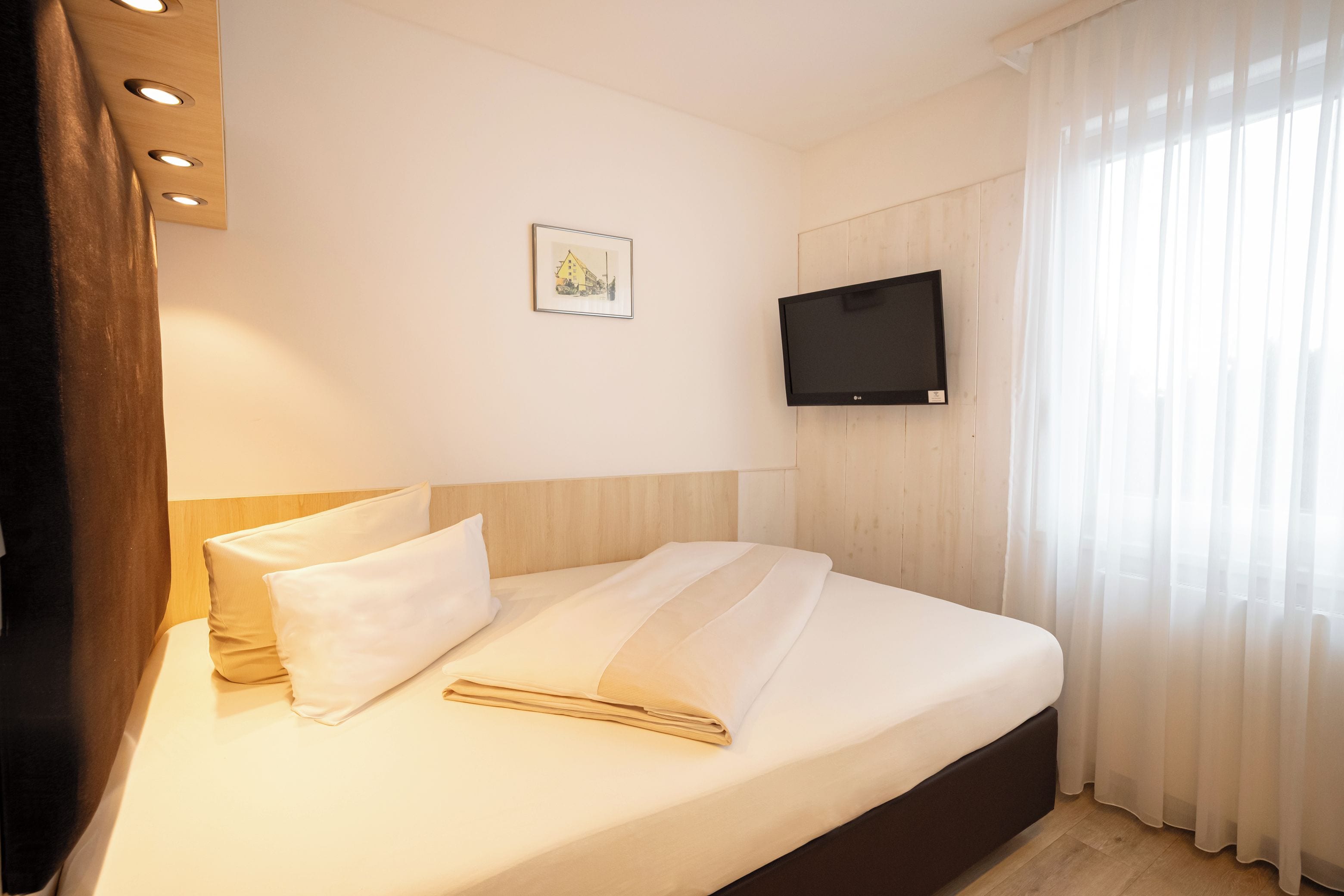 A hotel room with a bed, a flat-screen TV mounted on the wall, and a window with sheer curtains.