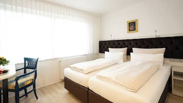 Ein Hotelzimmer mit einem Doppelbett, einem kleinen Tisch mit Stuhl und einem Fenster mit durchsichtigen Vorhängen.