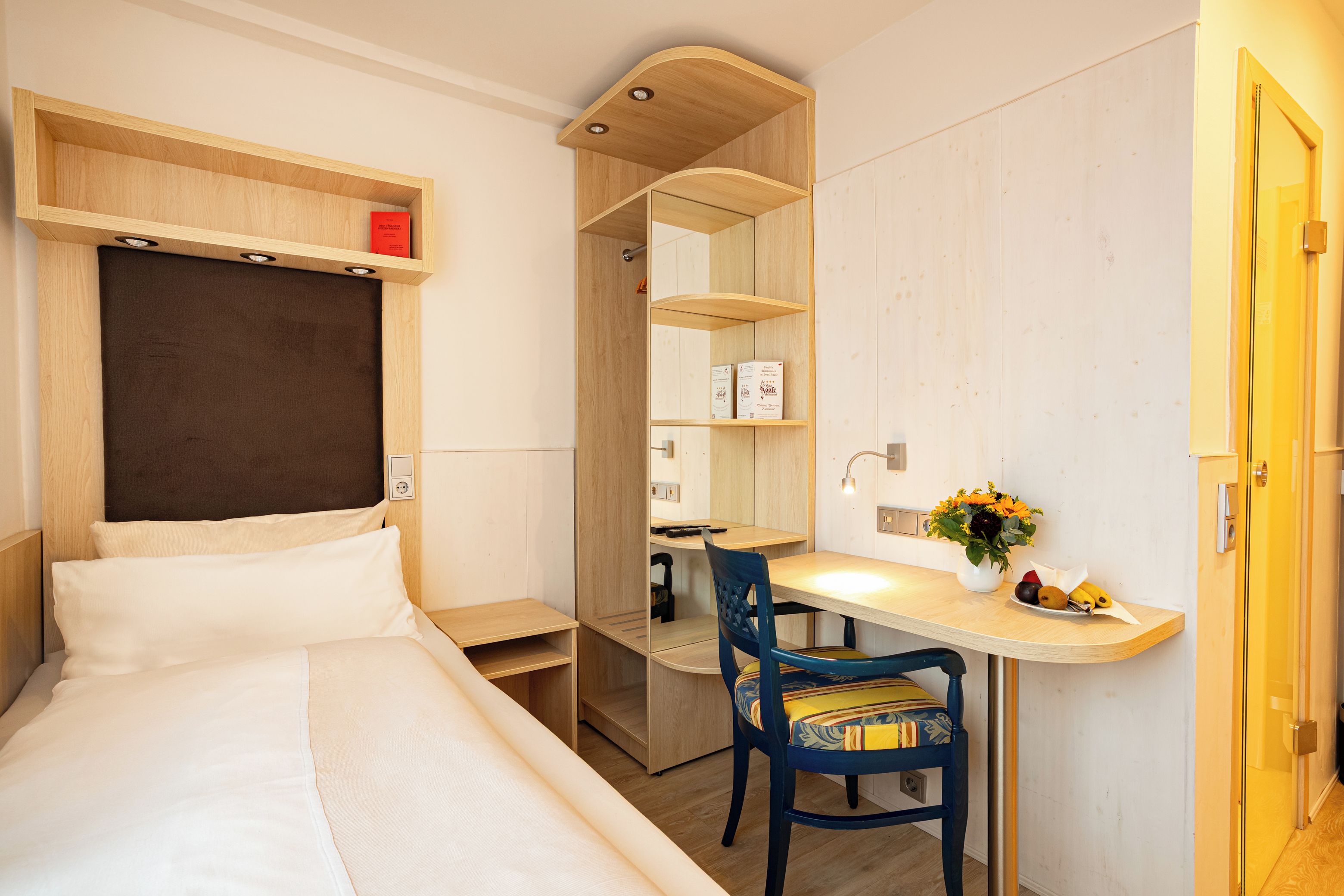 A hotel room with a bed, a desk, a chair, a mirror, and a small shelf with a red object.