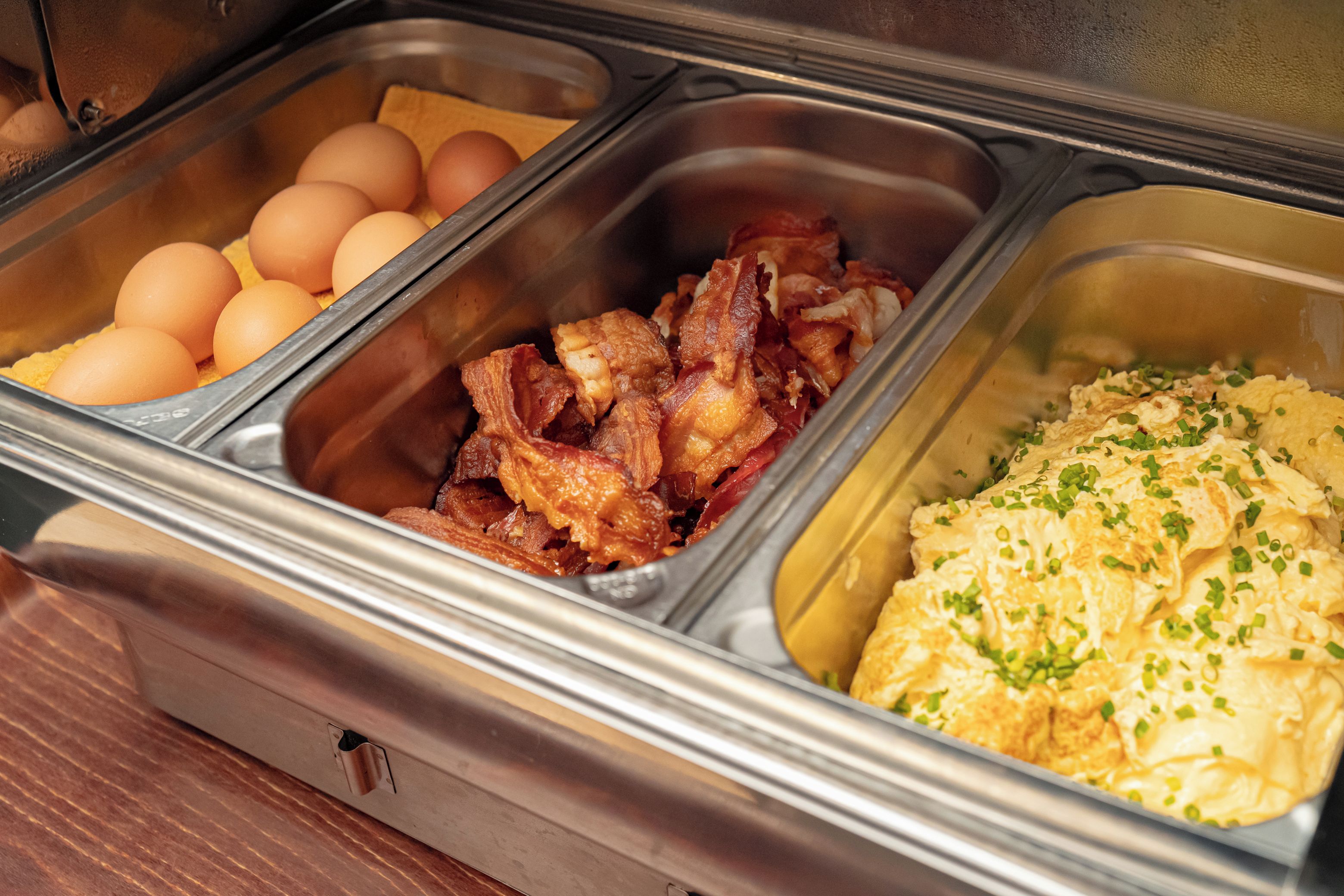 A buffet with labeled containers of eggs, bacon, and scrambled eggs.