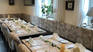 Een eetgedeelte met lange tafels gedekt voor bediening, een raam met een plant en een kroonluchter.