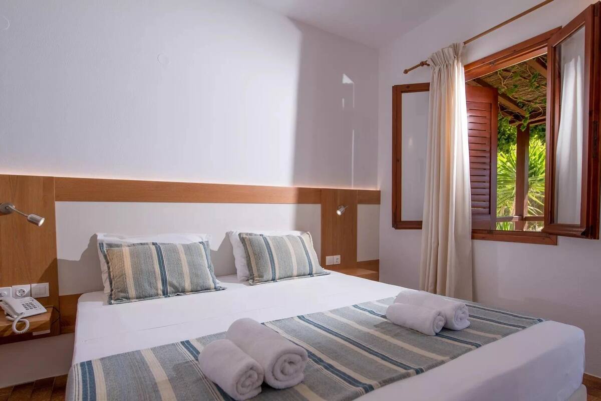 A hotel room with a bed, a bedside telephone, and a window with curtains.