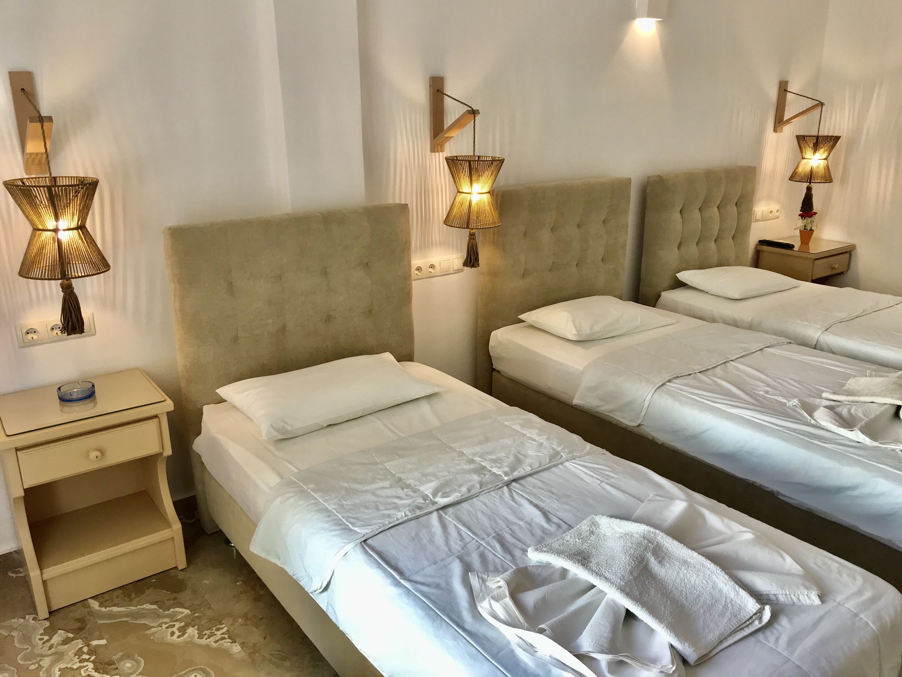 Habitación de hotel con dos camas, cada una con sábanas blancas y un edredón gris. Hay mesitas de noche con lámparas y un pequeño estante.