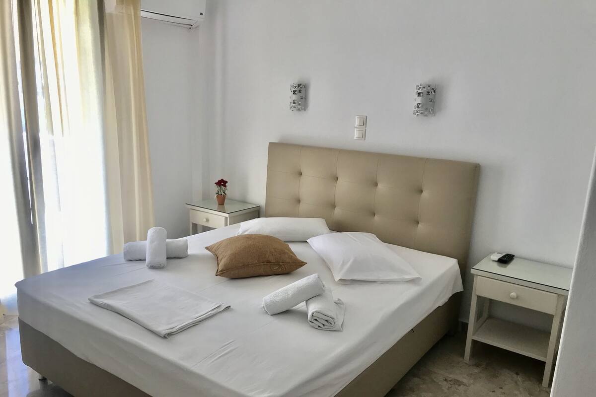 A neatly made bed with white linens, a beige headboard, and a bedside table with a remote control.