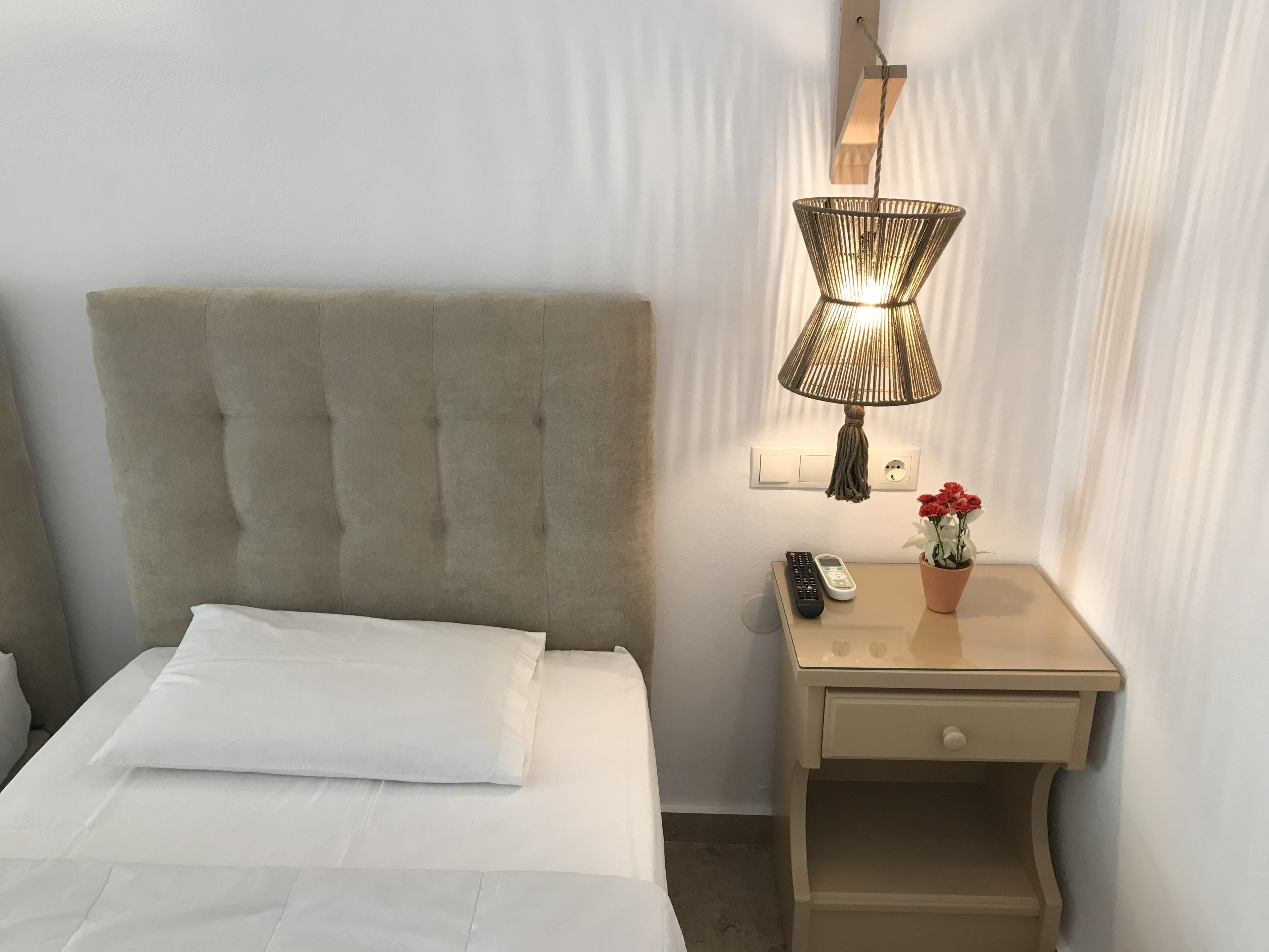 A neatly made bed with white linens, a tufted headboard, a bedside lamp, a remote control, a small potted plant with red flowers, and a nightstand drawer.