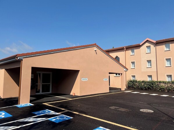 Een gebouw met receptie en kamers, een parkeerplaats met parkeerplaatsen voor mindervaliden, en een helderblauwe lucht.