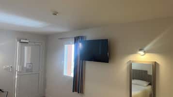 Habitación de hotel con cama, televisor montado en la pared, ventana con cortinas, puerta y mesita de noche con toallas.