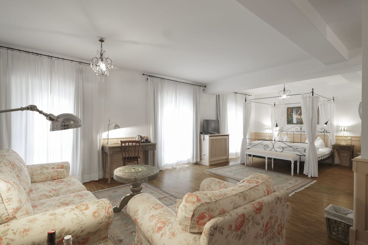 Een ruime woonkamer met sofabanken met bloemenprint, een ronde salontafel, een kroonluchter en een hemelbed met baldakijn.