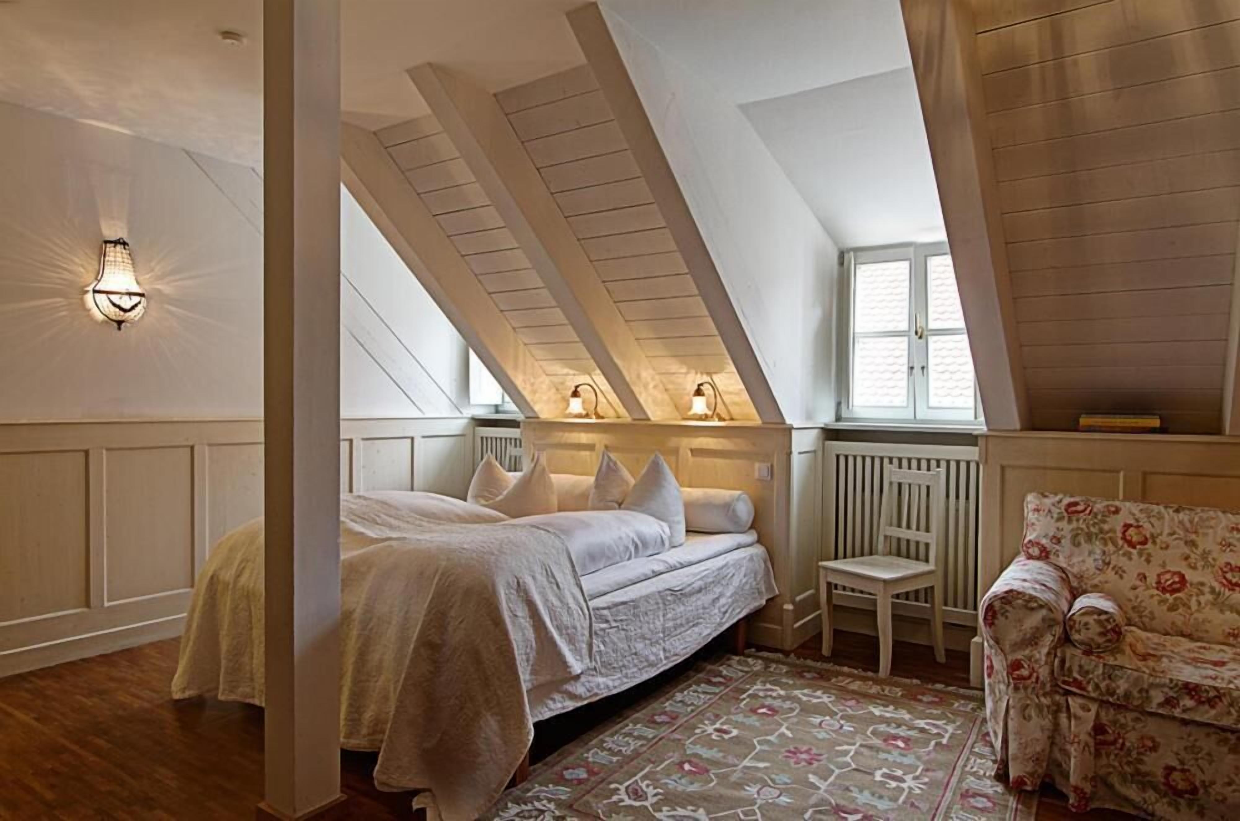 Een slaapkamer met een schuine kap, een bed met wit beddengoed, een stoel, een bloemenbank en een raam met daglicht.
