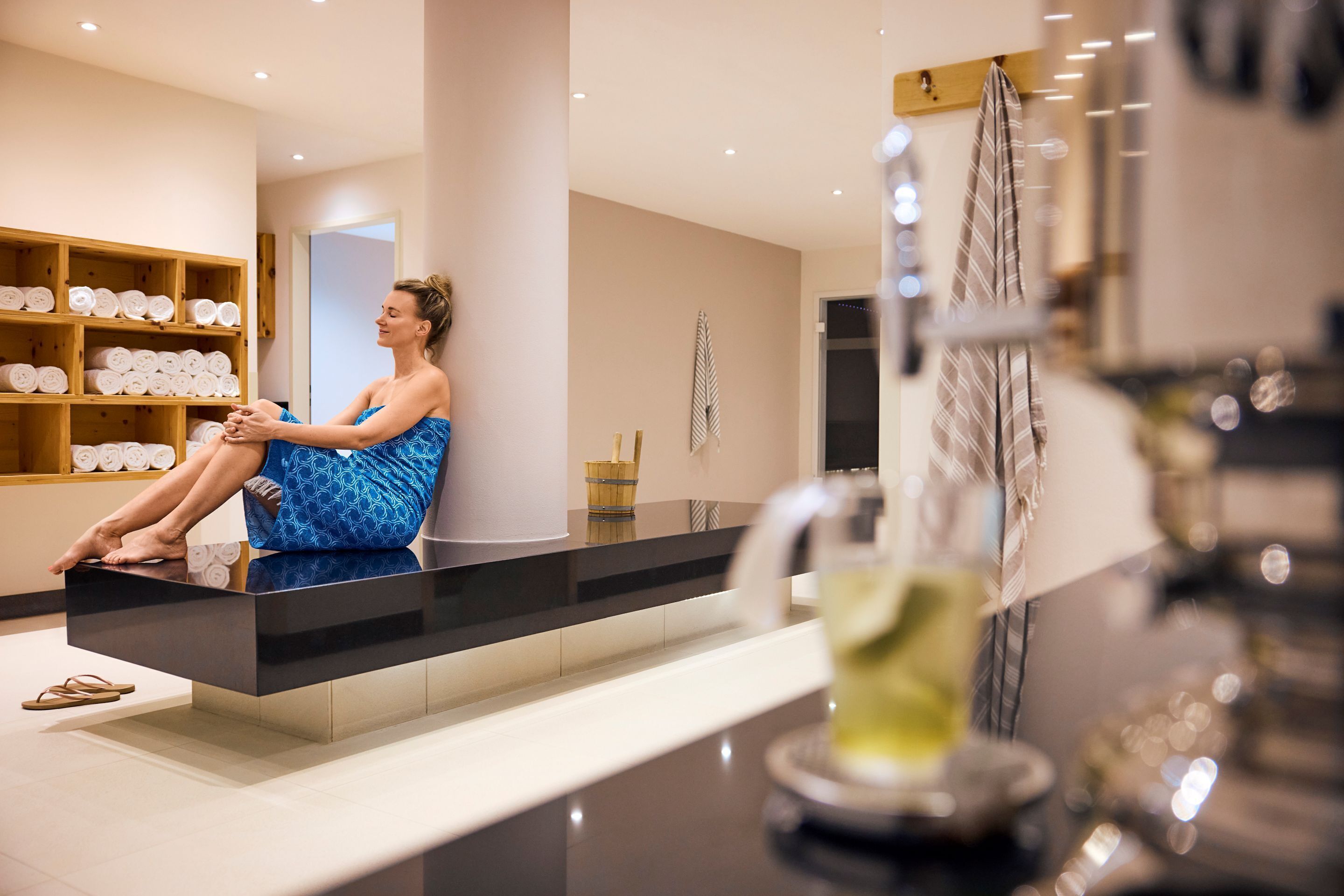 Una mujer sentada en un banco negro en un moderno espacio de spa, con estantes de toallas y un espejo grande.
