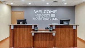 A reception desk with a "WELCOME THE ROCKY MOUNTAIN STATE" sign above. There are two computer monitors and a "Hilton Honors" sign on the desk.