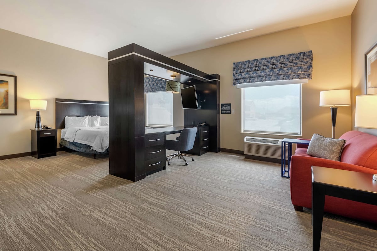 A hotel room with a bed, a desk, a chair, a red sofa, and a window with a view.