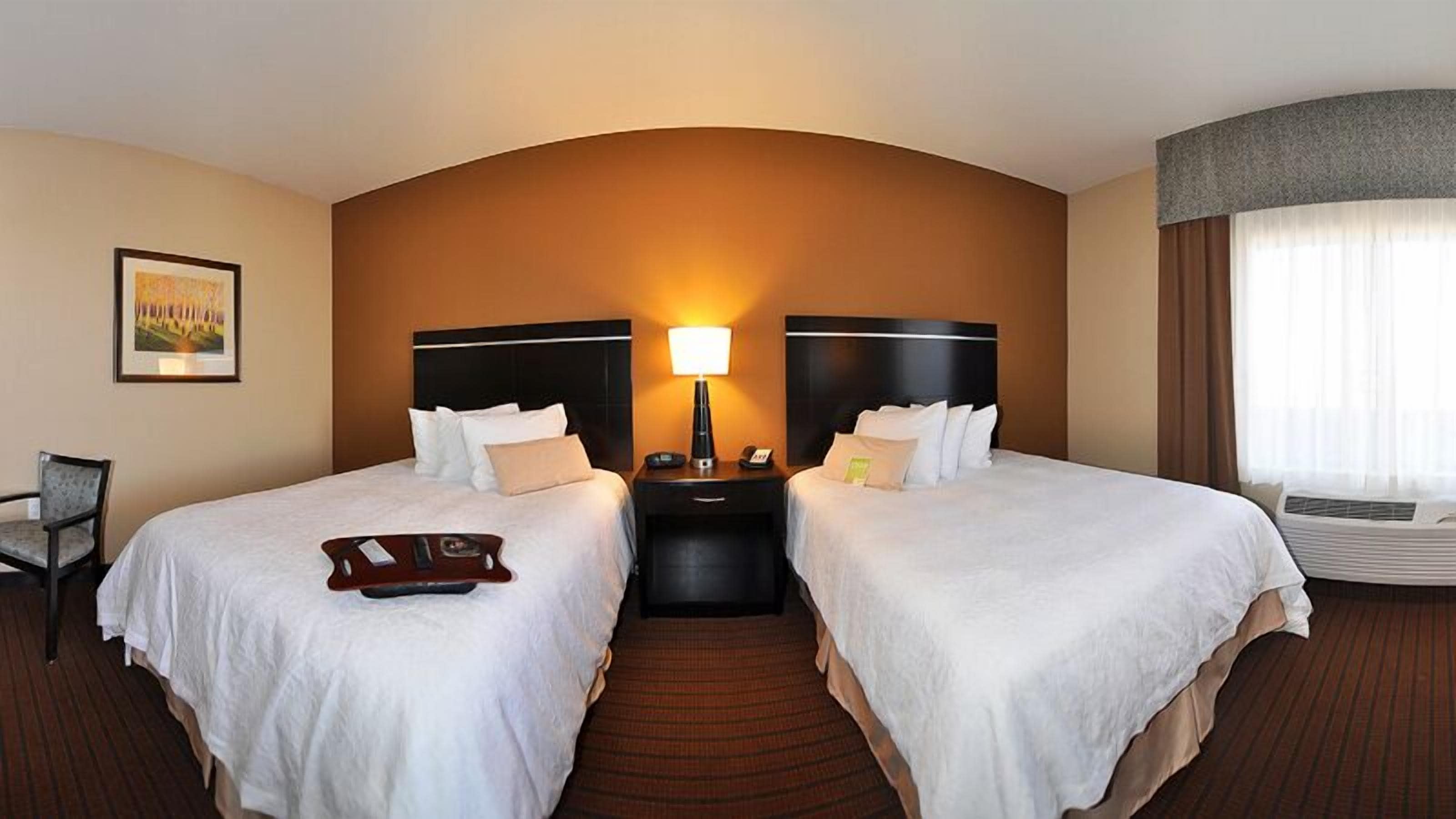 A hotel room with two beds, a nightstand with a lamp, a chair, and a large window with curtains.