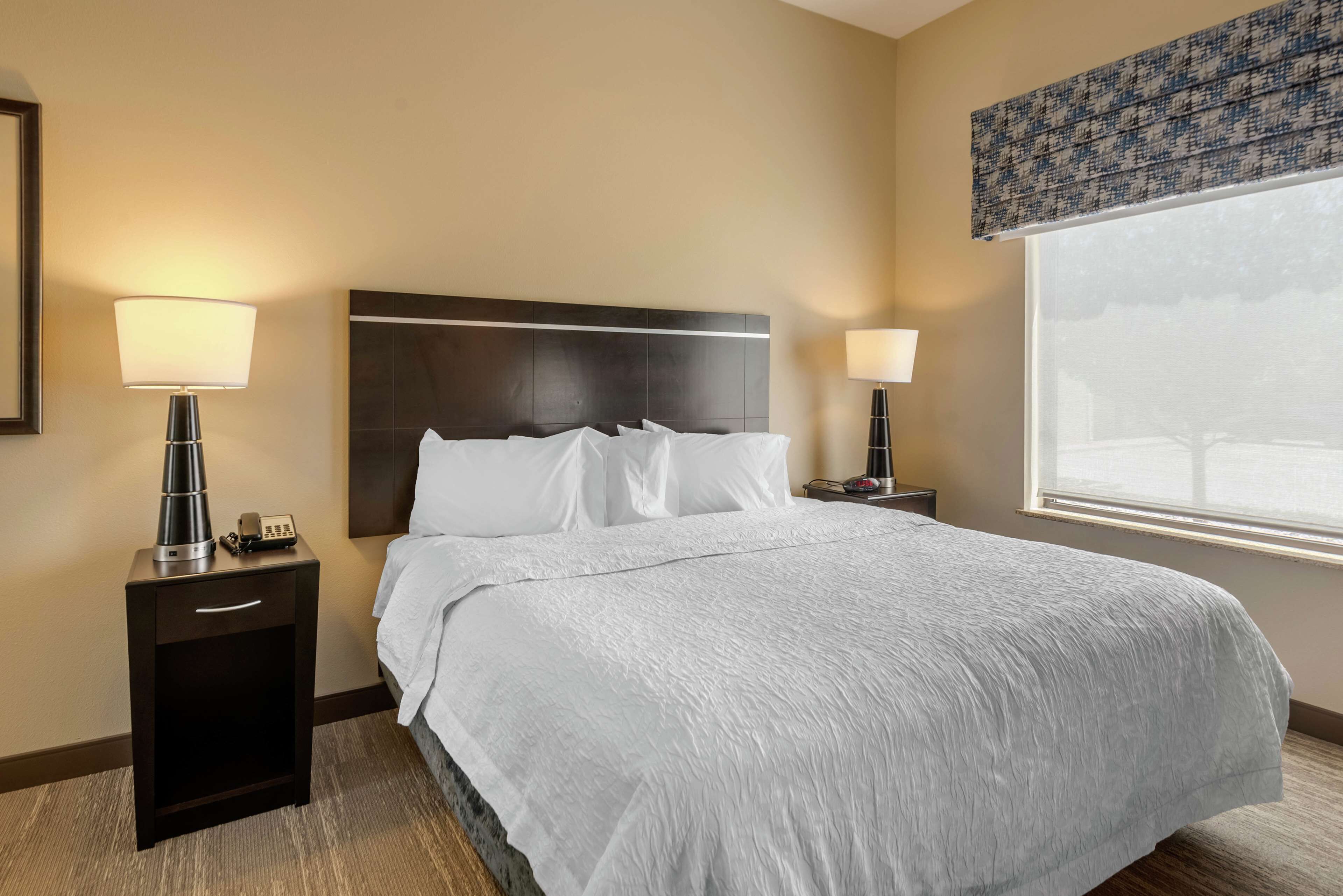 A hotel room with a large bed, two bedside tables with lamps, a nightstand, and a window with a view.