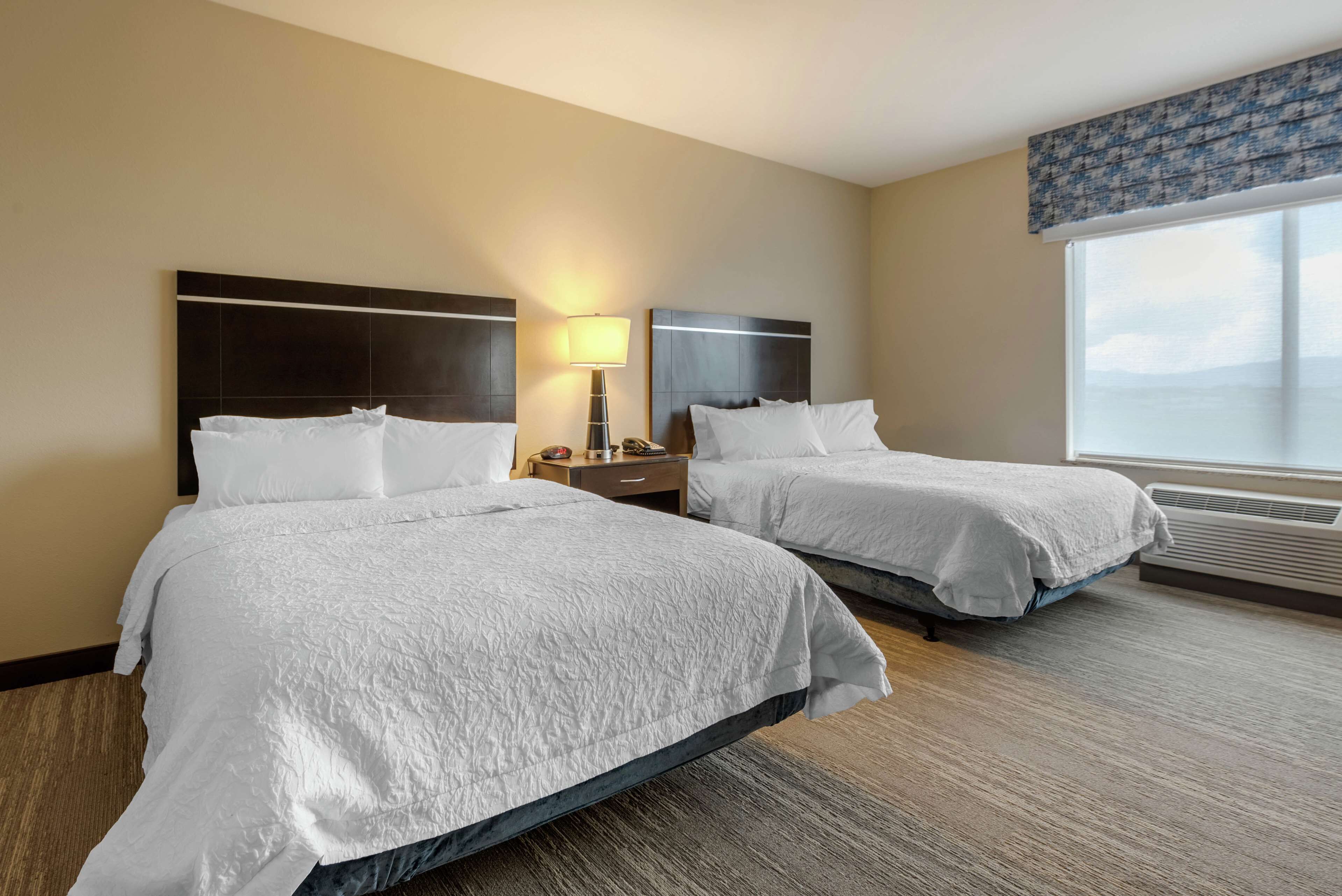 Two beds with white linens in a hotel room with a large window and a nightstand with a lamp.