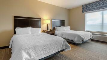 Two beds with white linens in a hotel room with a large window and a nightstand with a lamp.