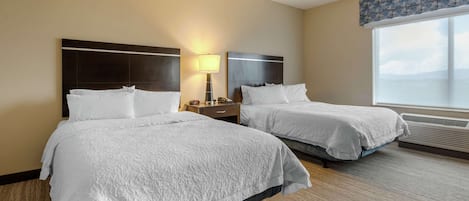 Two beds with white linens in a hotel room with a large window and a nightstand with a lamp.