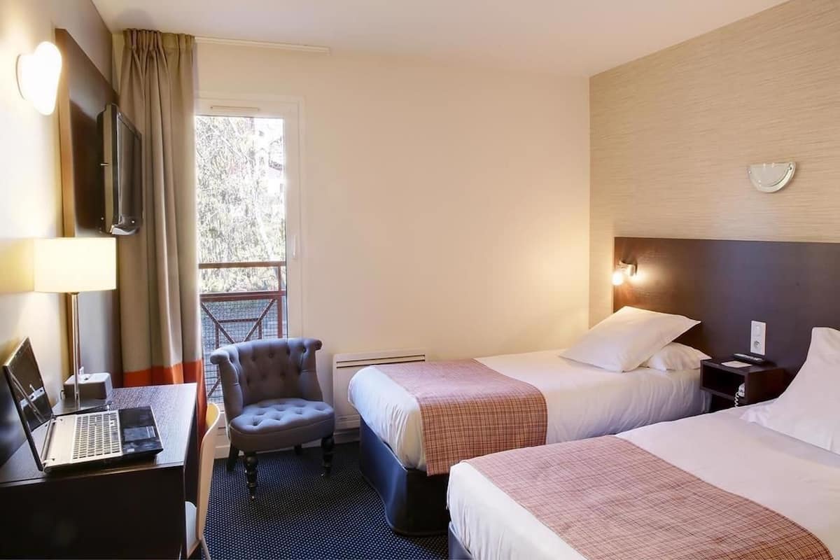 Habitación de hotel con dos camas, un escritorio con una computadora portátil, una silla, un televisor y una ventana con cortinas.