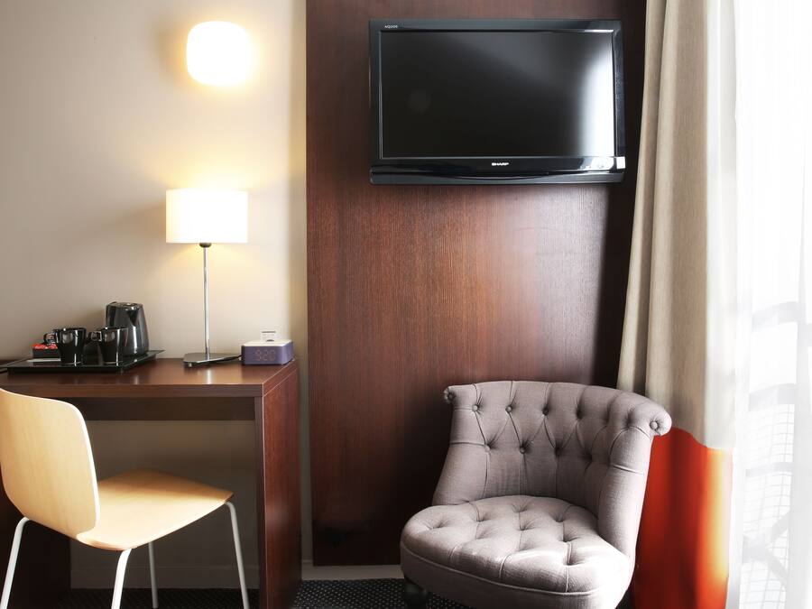 Habitación de hotel con televisor de pantalla plana montado en la pared, un escritorio con lámpara, una silla y una cafetera.