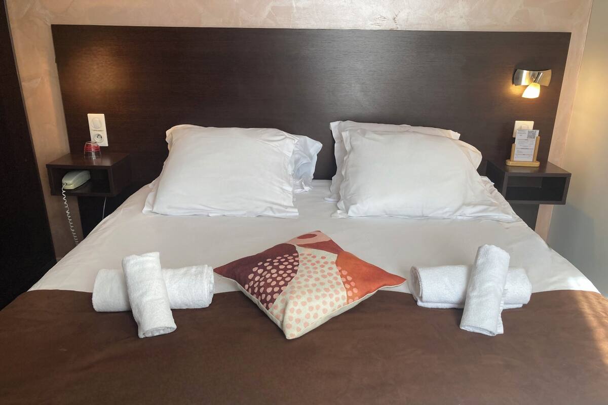 A neatly made bed with white linens, a polka-dotted pillow, and rolled towels on a brown bedspread.