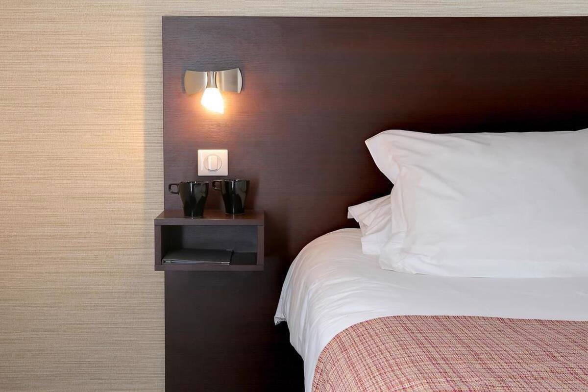 A hotel room with a bed, a nightstand with a lamp, and a wall-mounted shelf.