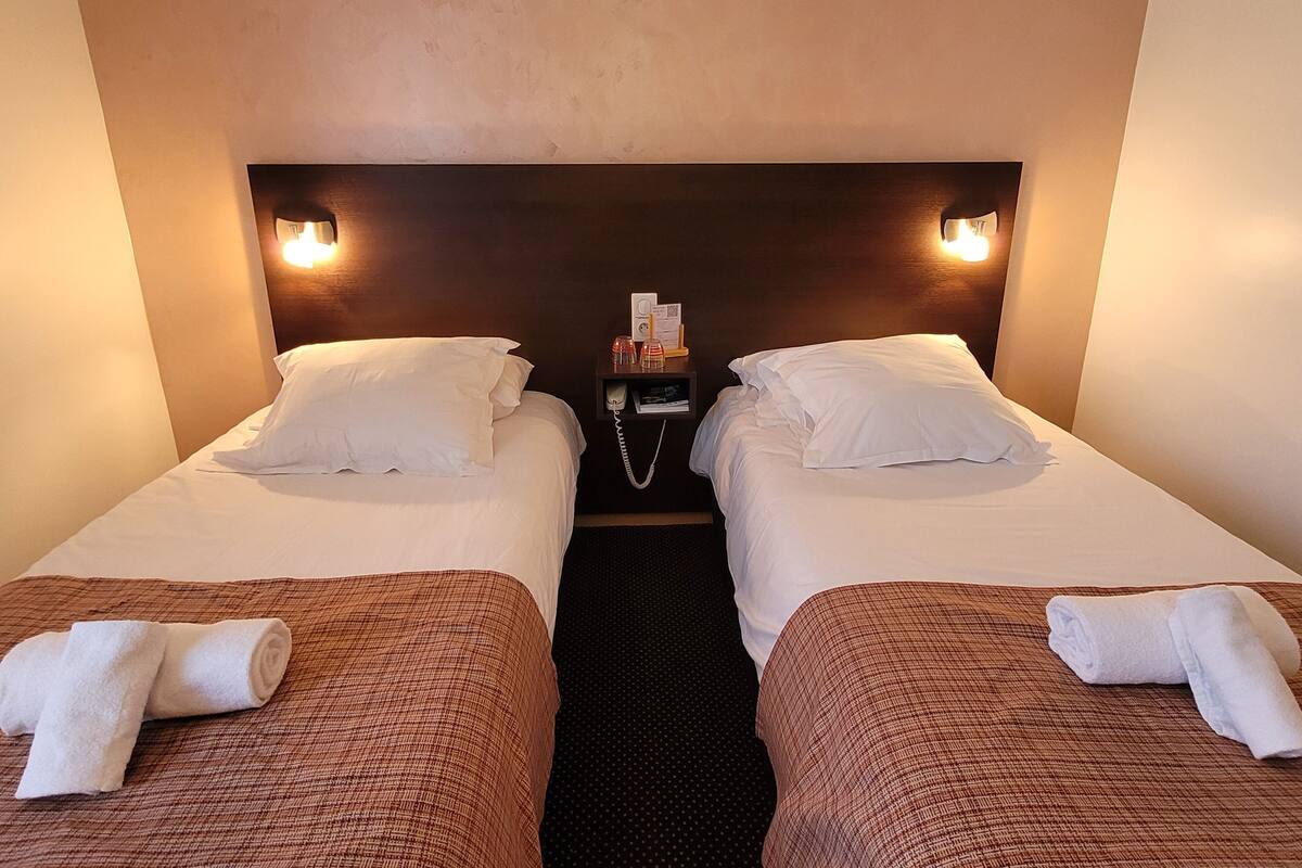 A hotel room with two single beds, each with white linens and pillows. There is a bedside table with a telephone and a small tray of toiletries. The room has a simple, clean design with a dark headboard and wall sconces providing light.