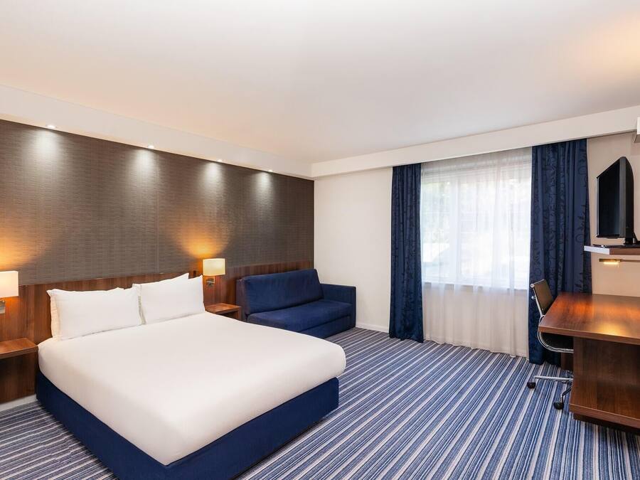 Quarto de hotel com uma cama grande, duas mesas de cabeceira com luminárias, um sofá azul, uma TV sobre um suporte de madeira e uma janela com cortinas.