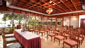 A spacious indoor area with a long table and multiple chairs, wooden ceiling, and large windows with a view of palm trees. - Coco Bay Resort (Kottayam)