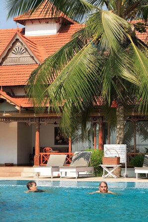 A pool area with lounge chairs, a palm tree, and a building with a tiled roof. - Coco Bay Resort (Kottayam)