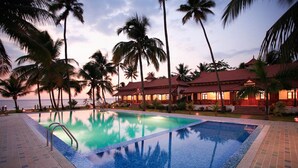 A swimming pool with a tiled deck, surrounded by palm trees and a building with a red roof. - Coco Bay Resort (Kottayam)