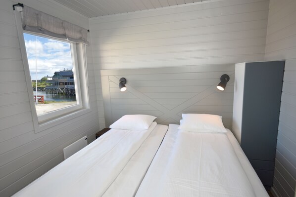 A room with two beds, a window with a view of a dock and buildings, and a wall-mounted cabinet. - Lankanholmen Sea Cabins (Andoy)