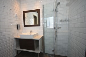 Un baño moderno con azulejos blancos tipo metro, un lavamanos blanco, un mueble de tocador de tonos oscuros, un box de ducha de vidrio y un espejo con marco de madera.