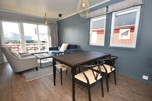 Una sala de estar moderna con piso de madera, un sofá, una mesa de comedor con sillas y ventanas grandes con cortinas.